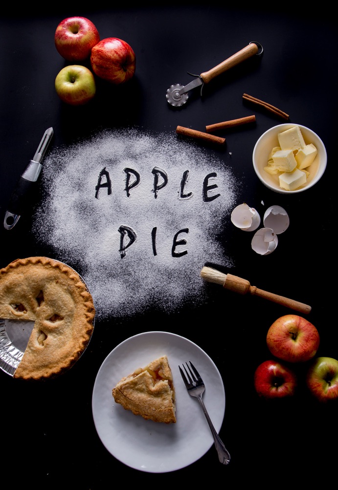 Overhead baker's table with APPLE PIE written in flour, surrounded by pie, apples, butter and cinnamon