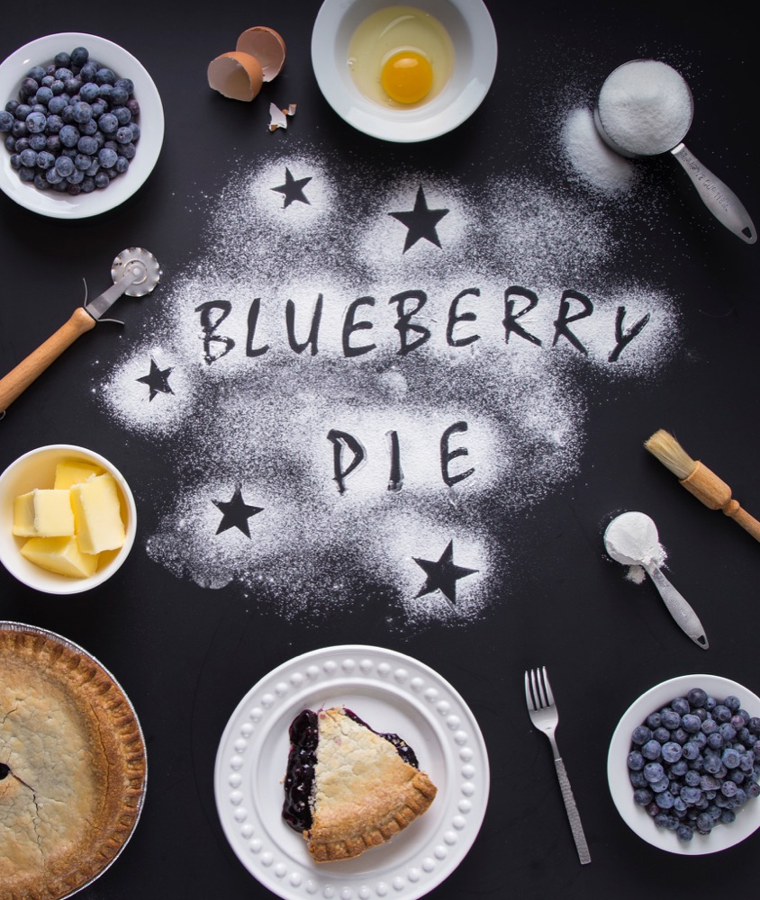 Overhead baker's table with BLUEBERRY PIE written in flour, with bowls of blueberries, pie and butter