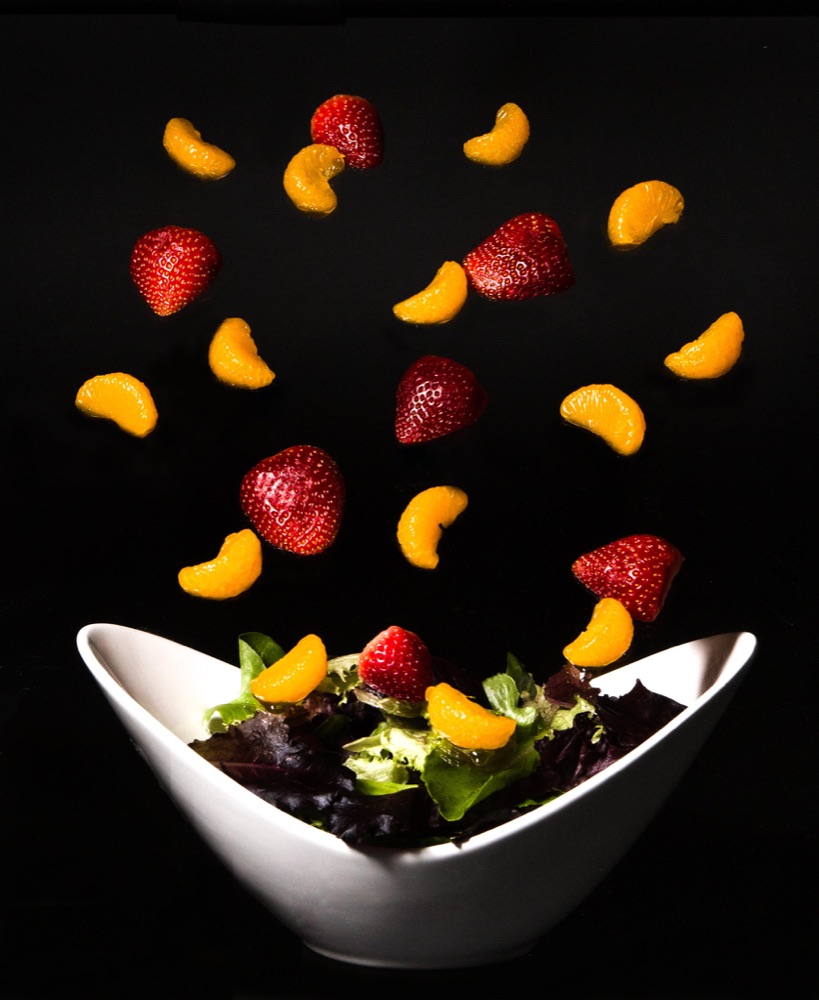 Strawberries and mandarin segments suspended in mid-air above a white bowl of greens, on black