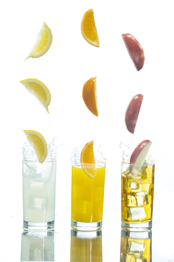 Three glasses of juice with lemon, orange and apple slices suspended in mid-air above them