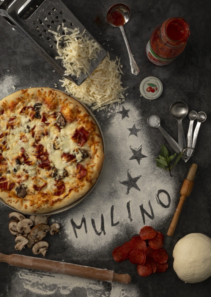 Rustic pizza scene with grated cheese, pepperoni, dough and the word Mulino written in flour