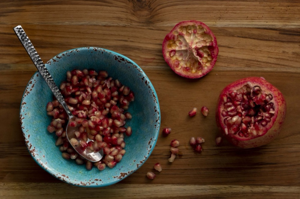 Pomegranate seeds in a turquoise bowl with a hammered spoon on warm wood