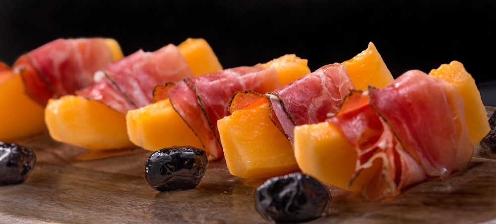 Prosciutto-wrapped melon wedges with black olives on a wooden board, against dark background