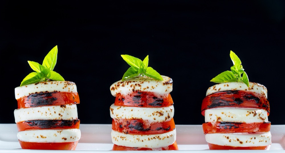 Three stacked towers of tomato and seared goat cheese topped with basil, on a white plate against black